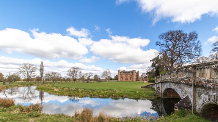 A view across the river Dene with the old stone bridge to the right, the main house in the far distance with the green grass of the paddocks stretching out in front of it. A blue sky with fluffy white clouds is above and reflected in the still water of the river.